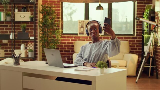 Young person having fun taking selfies at home office, working on online career with blogging. African american freelancer posing for photos with smartphone, self employed adult.