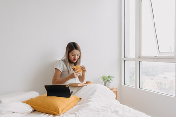 Asian Thai woman having breakfast, eating homemade sandwich while working at in apartment, using tablet in the morning time, busy at home.