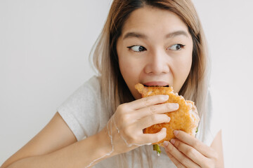Thai Asian woman eating and having breakfast, homemade sandwich hungrily with messy hands, looking at something while chewing bread, living alone in apartment isolated on white wall.