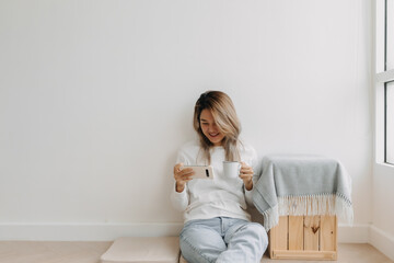Asian Thai woman enjoy using and holding smartphone while drinking coffee in apartment winter time. Happy moment smiling and chilling alone, sitting on floor next to window.