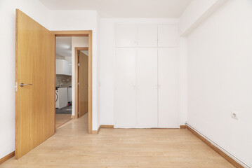 An empty room with a built-in wardrobe with white doors, freshly painted smooth walls, light-colored floors and an oak wooden door leading to a hallway