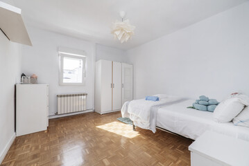 bed in a room with a white bedspread, some towels and cushions, a three-section wardrobe and a white dresser with drawers and plain white painted walls