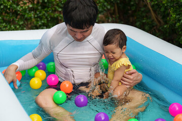 Fototapeta na wymiar happy father and infant baby girl playing water splashing with colorful plastic balls in inflatable pool