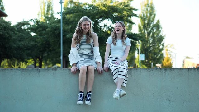 Two beautiful young women sit on a wall in a park and laugh