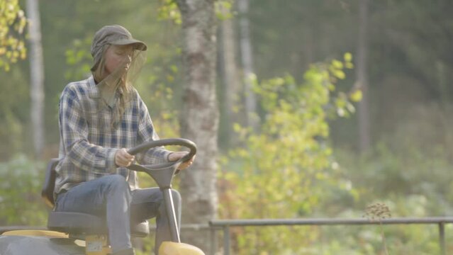 Lady on ride-on mower in countryside wearing mosquito head, rack focus