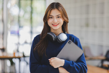 Young adult happy smiling Hispanic Asian student wearing headphones standing indoor using digital tablet in university campus or at virtual office. College female student learning remotely.