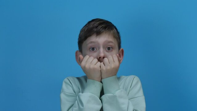A little Caucasian boy looks at the camera, is surprised and covers his mouth with his hand.