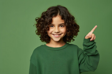 teenage girl stands against a green background in a studio, wearing a broad smile and pointing upward, expressing a wow emotion. advertisement concept, copy space