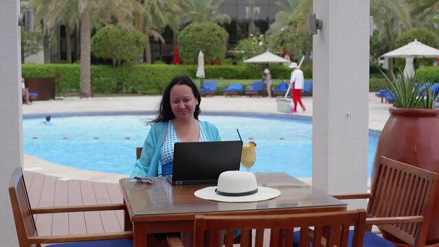 Happy woman stretching arms while using laptop. Freelancer in casuals sitting against swimming pool. She is relaxing in hotel during summer holiday.