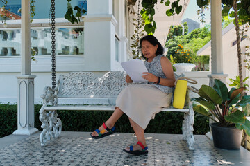 Female lecturer sitting on a garden bench in her house, reading an article on the printed paper