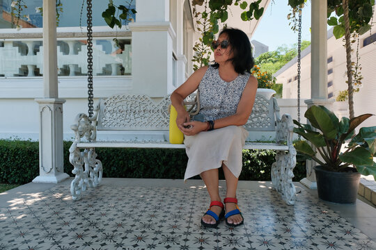 An Asian woman wearing sunglasses, sitting on a park bench in her home garden