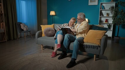 In the frame, an elderly couple sits on a couch in an apartment, against a blue wall. They are looking at, leafing through a photo album. They are chatting, reminiscing about old times, nostalgic