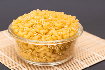 Uncooked Chifferi Rigati Pasta in Glass Jar on Bamboo Mat on Black Background. Fat and Unhealthy Food. Classic Dry Macaroni. Italian Culture and Cuisine. Raw Pasta