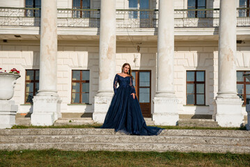 Naklejka premium Beautiful brunette woman with curly hair and red lips in long blue dress walking near big white palace with columns. Princess in palace conception