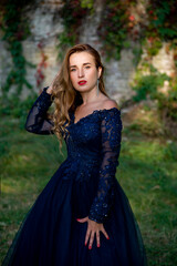 Fototapeta premium Beautiful brunette woman with curly hair and red lips in long blue dress walking in the park