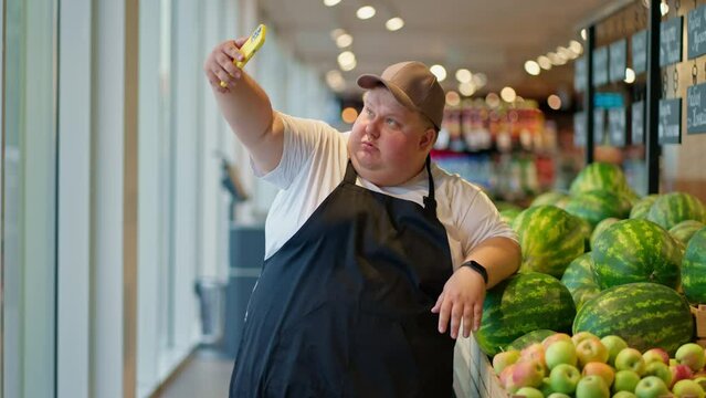 A funny and happy overweight male supermarket worker in a white T-shirt and a black apron in a gray cap takes a selfie using a yellow phone against the background of watermelons in the supermarket