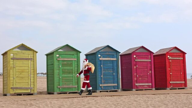 Santa Claus walks to the beach past the multi-colored beach huts. Christmas or New Year holidays in hot countries. Santa's summer vacation.