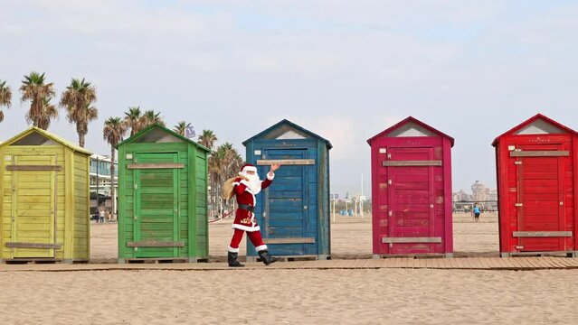 Santa Claus walks to the beach past the multi-colored beach huts. Christmas or New Year holidays in hot countries. Santa's summer vacation.