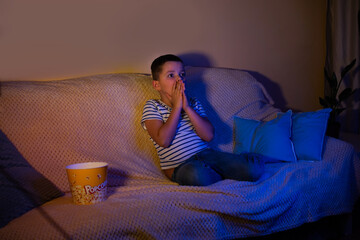 A little boy covered his mouth with his hands in horror and with big eyes watches a movie in a dark...