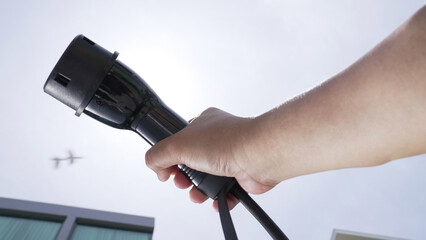 Closeup hand grasping an EV plug for electric vehicle with the midday sky in the background as progressive idea of alternative sustainable clean and green energy for environmental concern.