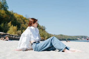 Fototapeta na wymiar Side view of a carefree young slender woman in casual clothes sitting on seashore, relaxing on a sunny day at sea
