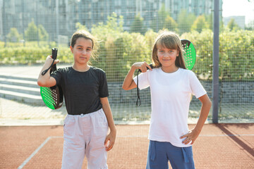 Obraz premium Kids and sports concept. Portrait of smiling girls posing outdoor on padel court with rackets and tennis balls