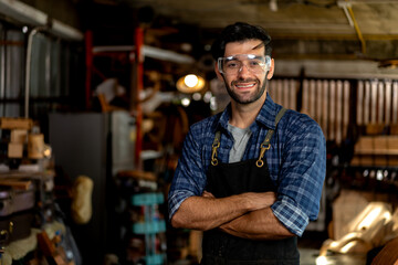 Portrait of a young handsome carpenter wearing a plaid shirt and apron, smiling in his workshop.