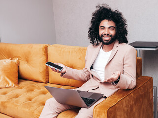 Handsome smiling hipster model. man dressed in elegant beige suit . Fashion male with long curly hairstyle posing at home. sitting on yellow sofa. Holding laptop, using notebook and smartphone, phone