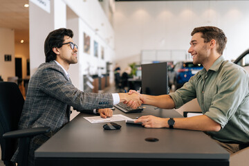 Side view of happy smiling client male purchasing automobile in dealership, signing paper, shaking hands with car dealer in suit enjoying successful agreement. Concept of buying new auto at showroom.
