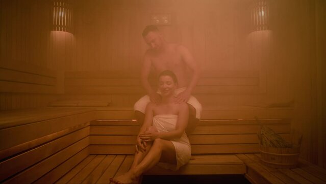 A married couple spends time in a Finnish sauna. The sauna is filled with steam. The man and the women are covered with towels. He massages her shoulders while she tells him something and smiles.