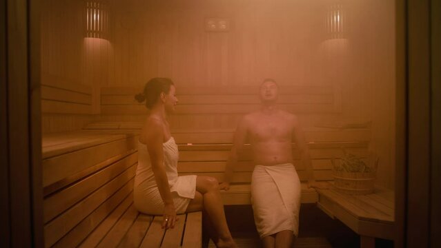 A woman and a man are sitting in a Finnish sauna. They are both covered with towels. The man waves a whisk over himself and puts it in a bucket. The woman says something to the man and he replies.
