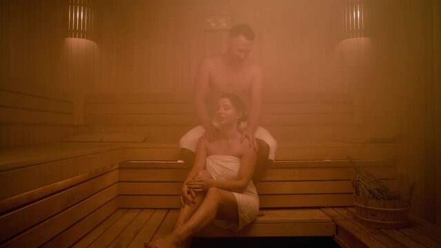 A woman receives a shoulder massage from her boyfriend. They are relaxing in a Finnish sauna. They are wearing towels and sweating. There is a whisk in a bucket nearby. The sauna is filled with steam.