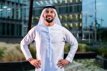 Poster Arab middle-eastern man wearing emirati kandora traditional clothing in the city - Arabian muslim businessman strolling in urban business centre. © oneinchpunch