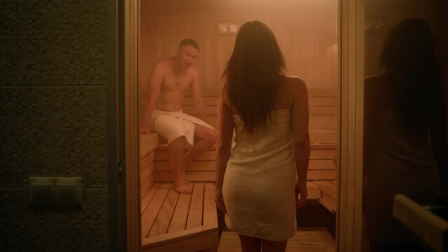 A gorgeous woman wrapped in a towel enters a steamy Finnish sauna. A man is already relaxing on a second floor bench. The woman sits on a first floor bench and relaxes her legs on it. A couple talks.