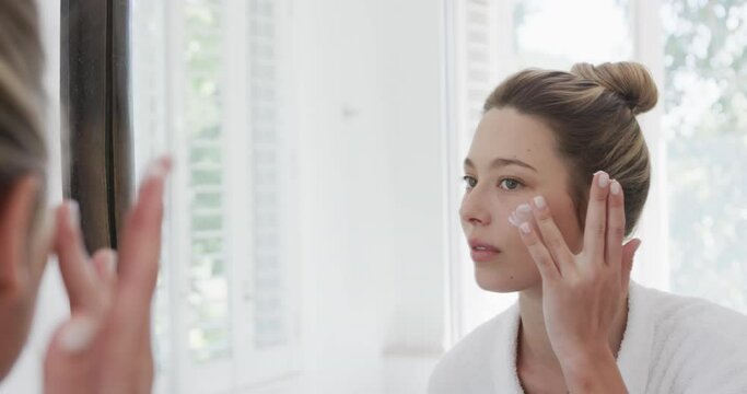 Biracial woman applying face cream looking at mirror in bathroom, slow motion