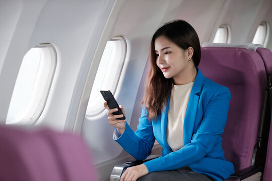 Young Asian Woman Sitting On Seat In Airplane And Talking By Smartphone. Young Woman Traveller Talking On Cell Phone