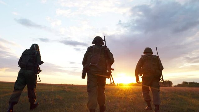 Silhouettes of three armed and fully equipped soldiers walking in field at sunset. Special forces squad in military service, war patrol operation and fight with weapon conflict