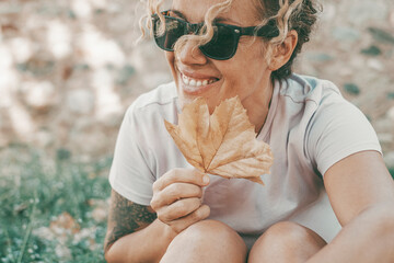 Cheerful outdoor portrait of happy young mature woman smiling and holding big yellow autumn maple leaf near the face. Concept of relax and outdoor leisure activity in the garden. Natural lifestyle