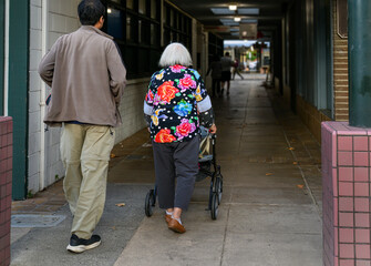Senior woman walking using a mobility walker in a shopping alleyway, accompanied by her son....