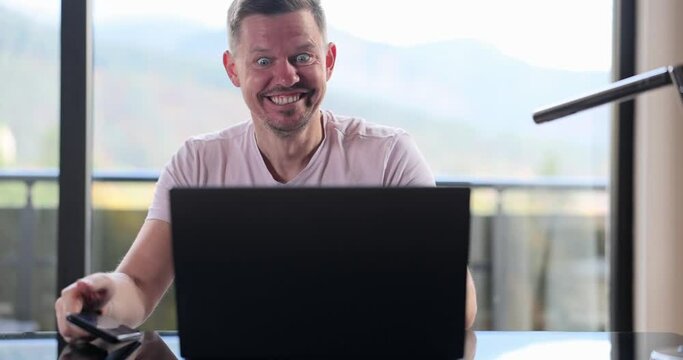 Man sits at table, reads email on smartphone and on laptop makes yes gesture and feels happy. Businessman receives great business news and celebrates career growth
