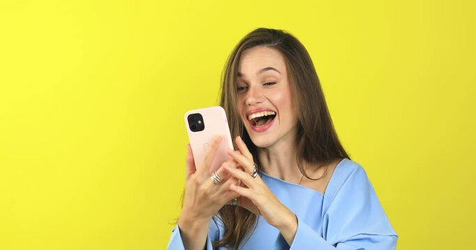 Attractive brunette curly woman look surprised wow hold using mobile cell phone on yellow background. Girl look on smartphone and doing winner gesture. Girl just found out big win news, win gesture.