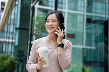 Portrait of beautiful asian woman holding mug and using smartphone with happiness while standing outside the office.
