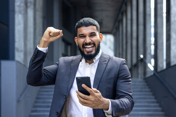 Portrait of a young Muslim businessman man standing near an office building, holding a phone in his hands and showing a victory gesture with his hand, looking happy at the camera
