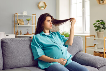 Portrait of a young brunette sad pregnant woman in casual clothes sitting on sofa in the living...