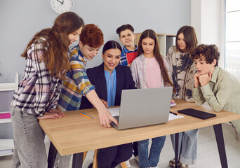 Happy group of children with female teacher using laptop together. Junior school students in casual wear gathered at teacher table looking at laptop screen in modern school classroom