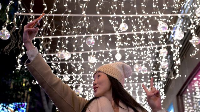 Young cheerful girl taking a selfie in the street at christmas night. Happy woman wearing warm hat taking picture with her phone in winter.