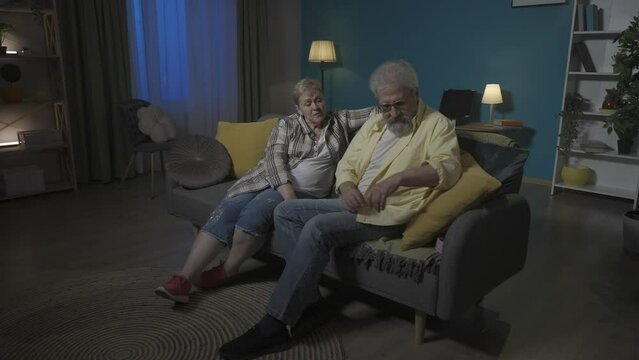 In the picture, an elderly couple is sitting on a sofa in a room. Men reach for a pink box hidden under the pillow. A gift for a woman, wants to surprise her, she does not see or know. HDR BT2020 HLG
