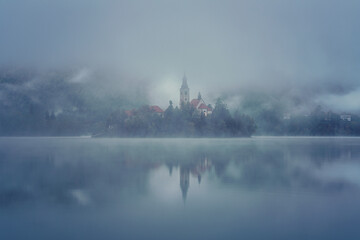 Famous alpine Bled lake (Blejsko jezero) in dense fog, amazing misty landscape, Slovenia. Scenic view of the lake, island with church and reflection in the water, outdoor travel background