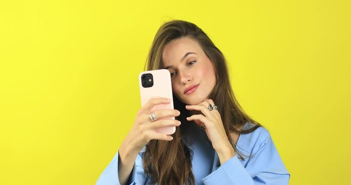 The girl stares at the phone and fixes her hair. Female uses phone like mirror. Young girl make photo from hands with phone on yellow background, look at camera and smiling.