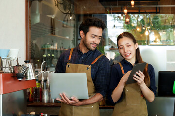 Young barista Asian woman and man of many nationalities business couple small cafe. Looking monthly income check from laptop and tablet. received large number coffee bakery customers happy smiles